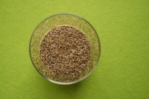 Cumin seeds in a clear glass bowl on a vibrant green background, viewed from above.