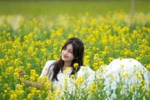 A young woman in a white dress smiles while relaxing in a vibrant mustard field during the day.